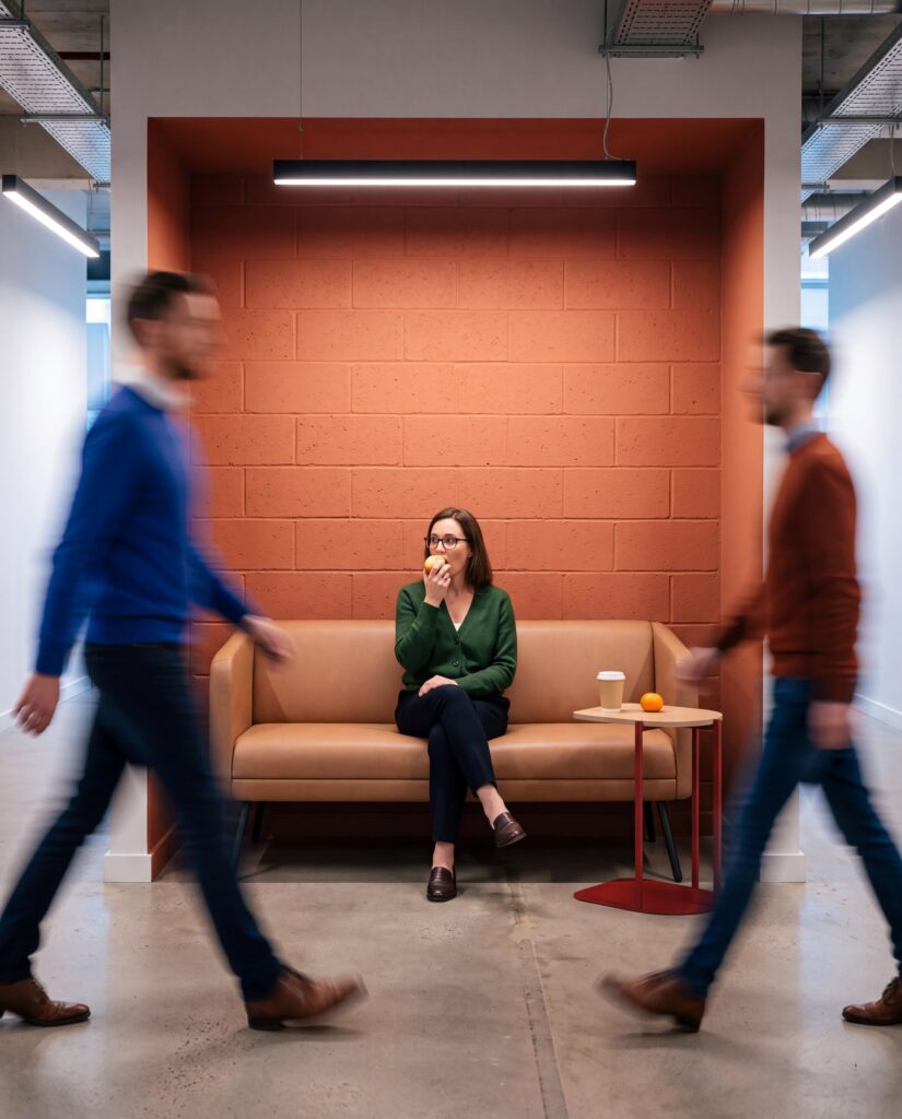 Set — Woman sitting on a beige leather waiting sofa in front of a terracotta brick wall, with a side table and two people walking by