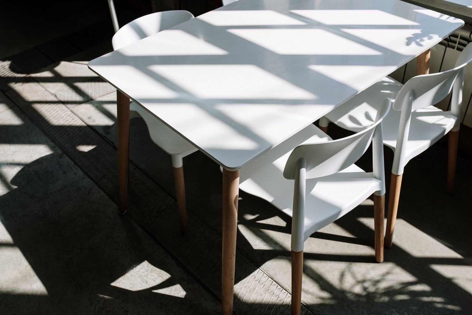A minimalist setup of white table and chairs with sunlight streaming through a window casting shadows indoors.